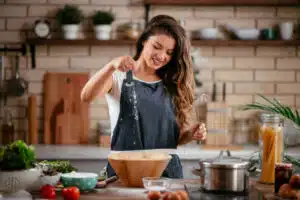 A woman in a kitchen preparing a homemade meal, sprinkling salt into a wooden bowl while smiling.