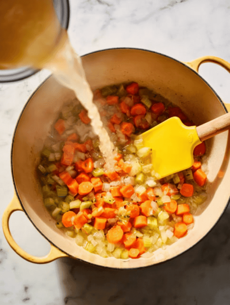 Pouring chicken broth into sautéed vegetables for lemon chicken orzo soup