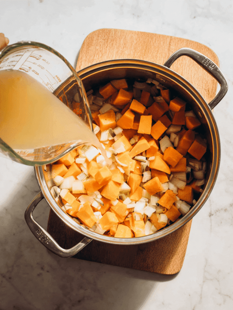 Pouring vegetable broth into a pot of butternut squash, onions, and apples to make butternut squash and apple soup with coconut milk.
